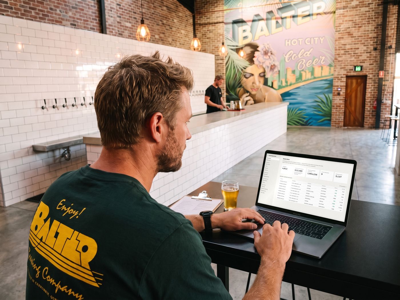 A Balter team member in the Enjoy! Balter tee at a high table in the taproom, the Club Enjoyment admin overview on his laptop, with the tap wall and the Hot City, Cold Beer mural beyond