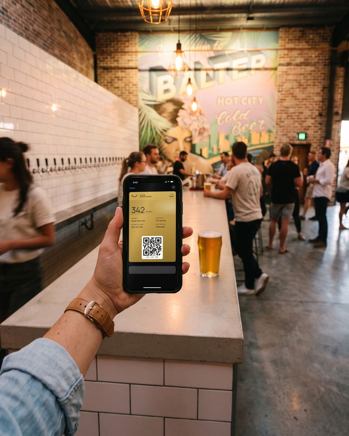 Sarah's Gold Club Enjoyment pass open in Apple Wallet at the bar on a busy evening, the mural glowing behind