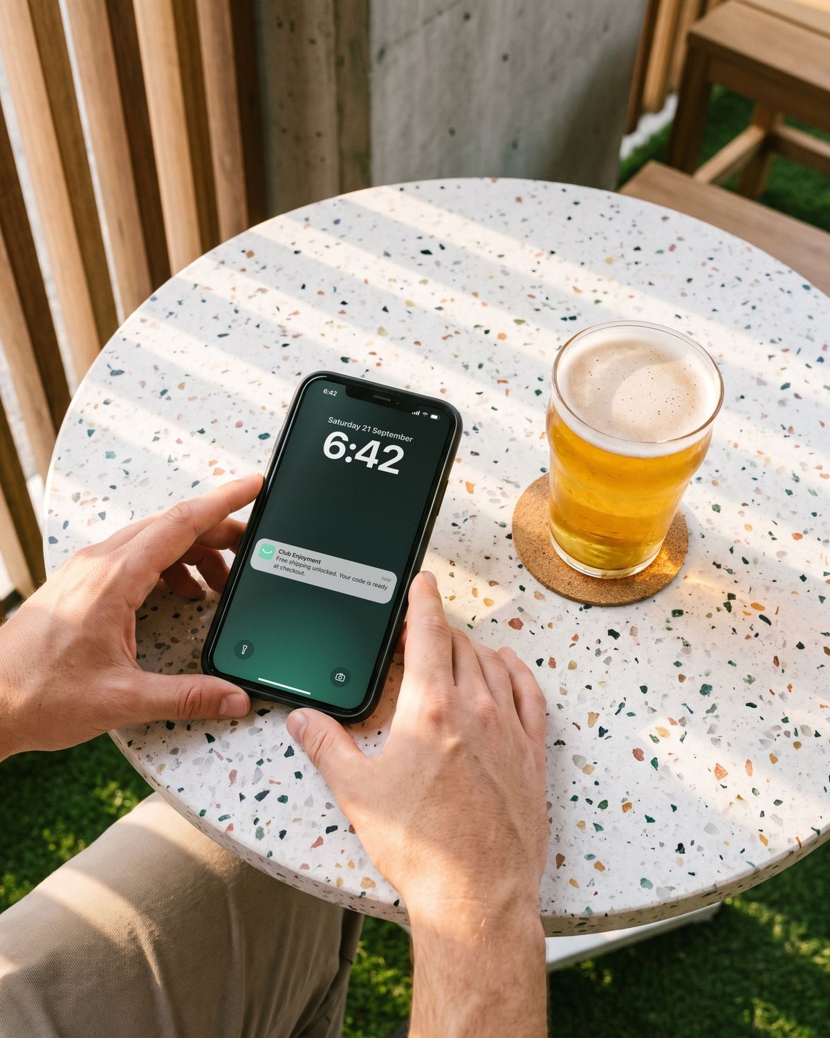 A phone on a terrazzo table in the beer garden beside a schooner, its lock screen showing a Club Enjoyment note: free shipping unlocked